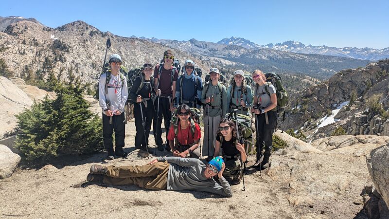A group of about a dozen people are posing for a photo on a rocky hilltop. They are surrounded by a mountainous landscape with sparse vegetation. Most of the people are wearing backpacks, suggesting they are hiking or backpacking. The sky is clear and blue, indicating a sunny day. One person is lying down in the foreground, adding a casual element to the group portrait.
